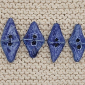 Può includere: Quattro bottoni a forma di diamante con una smaltatura blu profondo e lucida. Ogni bottone presenta una texture superficiale leggermente irregolare e organica, con due fori centrali per il fissaggio. I bottoni sono disposti in fila orizzontale, mostrando una finitura liscia e riflettente che mette in risalto le variazioni del colore blu sulle loro superfici.