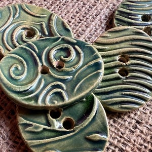 May include: A collection of handmade ceramic buttons in shades of green and brown. The buttons have a textured surface with a spiral or striped design and two or four holes for sewing. The buttons are oval-shaped and vary in size.