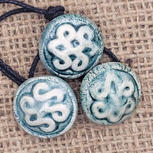 Op de afbeelding: Drie ronde, blauwgroene keramische hangers met witte, wervelende ontwerpen. Elke hanger is geregen aan een zwart koord. De hangers hebben een getextureerd, gecraqueleerd uiterlijk en worden gepresenteerd op een getextureerd, bruin oppervlak.