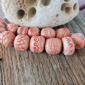 Może przedstawiać: Zbliżenie sznura różowych ceramicznych koralików z różnymi wytrawionymi wzorami. Koraliki są w kolorze jasnoróżowym z ciemniejszymi różowymi wytrawionymi wzorami. Koraliki są nawleczone na biały sznurek, na drewnianej powierzchni.