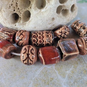 Può includere: Una collezione di perline in ceramica di varie forme e dimensioni. Le perline sono principalmente marroni e rosse, con intricati disegni intagliati in un marrone più chiaro. Alcune perline sono cilindriche, altre quadrate o ovali.