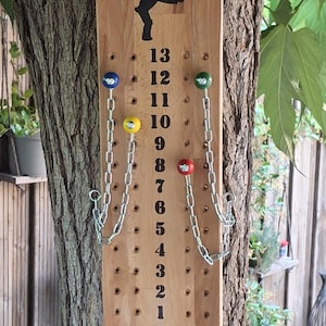 Può includere: Un tabellone segnapunti rettangolare in legno con la sagoma nera di una persona in alto e colonne verticali di numeri da 0 a 13. Include quattro pioli colorati attaccati a catene di metallo argentato per tenere traccia dei punteggi.