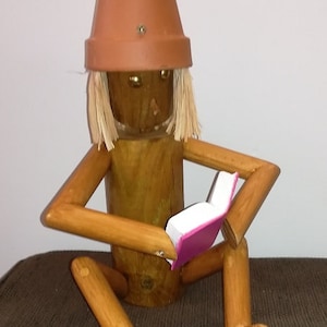 Può includere: Una statuetta di legno di una persona che indossa un vaso di terracotta come cappello e tiene un libro rosa. La statuetta ha i capelli di paglia ed è seduta su una superficie marrone.