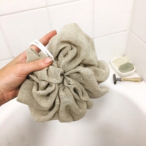 May include: A beige linen bath pouf with white drawstrings. The pouf is made of a soft, textured fabric and is designed for exfoliating and cleansing the skin.
