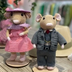 May include: Two crocheted mouse dolls, one wearing a pink dress and a purple hat, the other wearing a gray suit and a red tie.