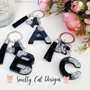 Può includere: Tre portachiavi in resina nera a forma di lettere con glitter argentato e nappe nere. Le lettere sono A, B e C. I portachiavi sono attaccati ad anelli portachiavi argentati. Il testo "Smelly Cat Designs" è visibile nella parte inferiore dell'immagine.