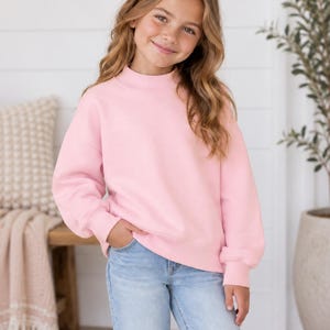 May include: A light pink long-sleeve sweatshirt featuring a mock neck collar, dropped shoulders, and ribbed cuffs at the wrists and hem, paired with light blue denim jeans. The sweatshirt has a relaxed fit and a solid, plain pastel pink color.