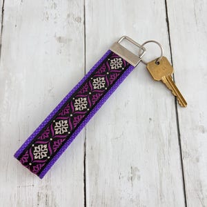 May include: A purple key fob with a decorative black and white pattern. The key fob has a silver metal clasp and a gold-colored key attached to it. The key fob is lying on a white wooden surface.