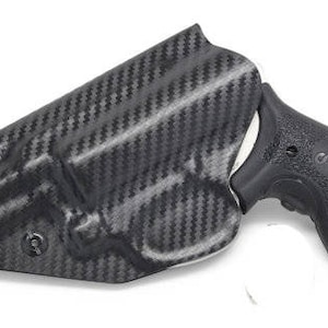 May include: A black carbon fiber holster with a textured pattern. The holster is designed to hold a black handgun grip. The grip has a textured surface and the letters "S&W" are visible.