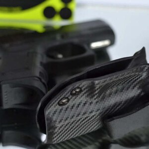 May include: Black handgun and a carbon fiber holster. The holster has a textured pattern and is designed to securely hold the firearm. The image is well-lit, showcasing the details of the gun and holster.