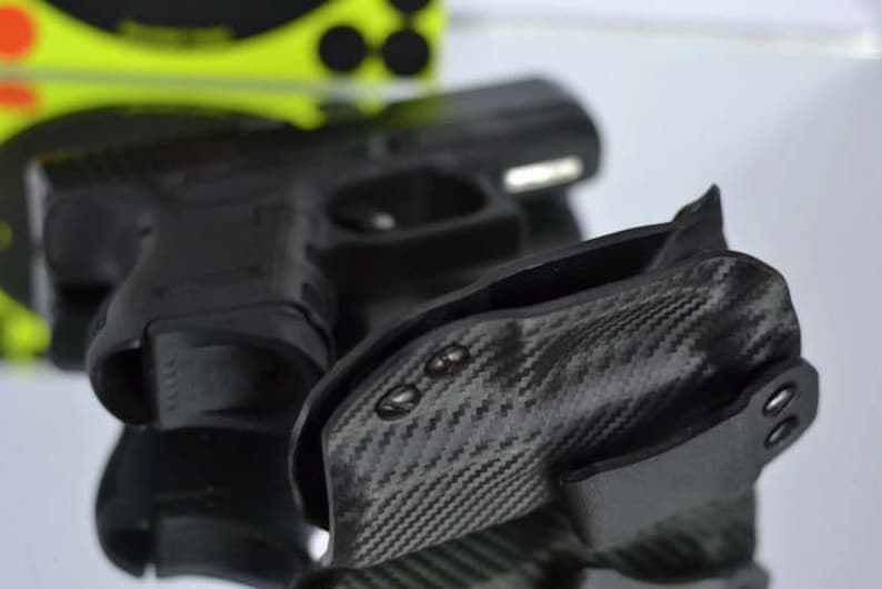 May include: Black handgun and a carbon fiber holster. The holster has a textured pattern and is designed to securely hold the firearm. The image is well-lit, showcasing the details of the gun and holster.