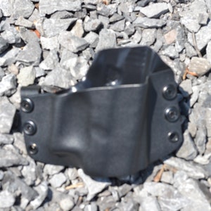 Puede incluir: Una funda de cuero negra con cuatro broches met&aacute;licos. La funda est&aacute; sobre una cama de grava gris.