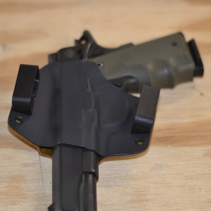 Puede incluir: Una funda de cuero negra con una pistola negra en su interior. La funda tiene dos clips en la parte posterior y un peque&ntilde;o lazo en el lateral.