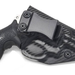 May include: A black handgun with a textured grip is partially inserted into a black and gray patterned holster. The holster has a belt clip and is secured with screws. The firearm is set against a white background.