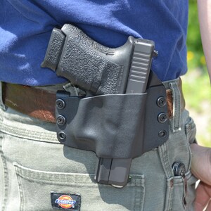 Puede incluir: Una pistola negra con una funda negra se lleva en la cadera de una persona. La funda est&aacute; asegurada a un cintur&oacute;n marr&oacute;n con tornillos negros. La persona lleva una camisa azul y pantalones verdes.