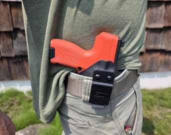Puede incluir: Una pistola naranja está enfundada dentro de la cintura, asegurada por una funda negra y un cinturón. La funda está sujeta a un cinturón de color caqui. La persona lleva una camisa verde y pantalones caqui.