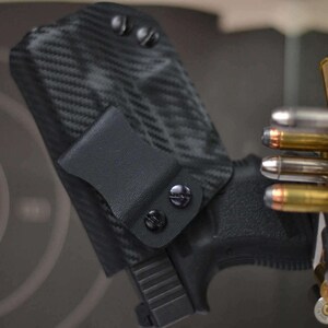May include: Black carbon fiber pattern holster with a black pistol. The holster has a belt clip and is secured with black screws. A collection of bullets is visible in the background, suggesting a shooting range or firearm-related theme.