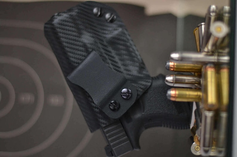 May include: Black carbon fiber pattern holster with a black pistol. The holster has a belt clip and is secured with black screws. A collection of bullets is visible in the background, suggesting a shooting range or firearm-related theme.