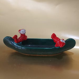 May include: A green ceramic canoe-shaped dish with two red figures with white faces on the sides.