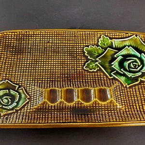 May include: A brown ceramic ashtray with a woven pattern and three indentations for cigarettes. The ashtray is decorated with green and white roses.