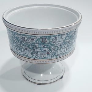 May include: A white ceramic bowl with a pedestal base. The bowl's exterior features a teal and white world map design. The rim and base have gold accents. The bowl is likely used for decorative purposes or serving.