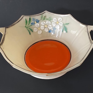 May include: A white ceramic bowl with two handles and a floral design. The bowl has a scalloped edge and a raised base. The center of the bowl is painted with a bright orange circle.