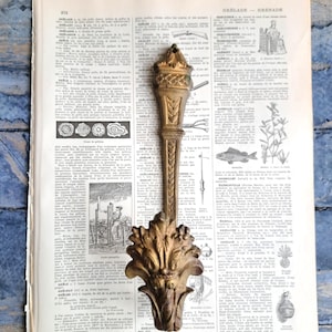 Könnte beinhalten: Verziertes, antikes, goldfarbenes Dekorationselement mit detailliertem floralem Design. Das Stück ist auf einer Vintage-Wörterbuchseite platziert, was einen Hauch von Geschichte und Eleganz verleiht. Die Oberseite hat ein fackelförmiges Design.