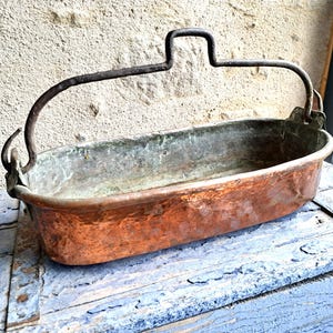 Op de afbeelding: Een antiek koperen bekken met een donker metalen handvat. Het bekken heeft een langwerpige vorm en vertoont tekenen van ouderdom met een gepatineerde afwerking. Het handvat is gebogen en aan de zijkanten van het bekken bevestigd. Het koper heeft een roodbruine tint.