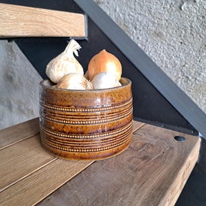 May include: A brown ceramic container with horizontal etched lines and filled with garlic and an onion. The container sits on a wooden surface, with a staircase and a concrete wall in the background. The container is a kitchen storage item.