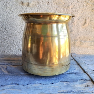 Op de afbeelding: Een vintage messing pot met een brede, uitlopende rand en een cilindrisch lichaam. De pot heeft horizontale geëtste lijnen rondom de omtrek. Het messing heeft een warme, gouden tint die het licht reflecteert. Het staat op een verweerd, blauw geschilderd houten oppervlak.