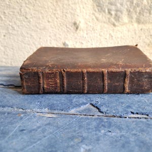 May include: A vintage, brown leather-bound Bible. The book's spine features the title "BIBLE" in gold lettering. The aged book is resting on a blue, weathered wooden surface, suggesting a historical or antique item.