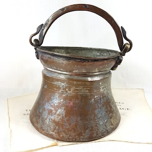 Puede incluir: Un cubo de cobre antiguo con un acabado desgastado y un asa curva con detalles decorativos. El cubo tiene una base ancha y una parte superior estrecha.