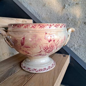 May include: A cream ceramic serving bowl with a red floral pattern. The bowl has two handles and a pedestal base. The rim and base are decorated with a floral border. The bowl is likely of French origin.