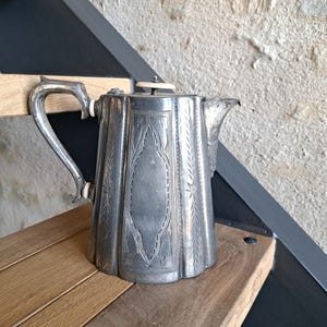 Puede incluir: Una cafetera antigua de color plateado con diseños grabados. La cafetera tiene un asa curva y un pico vertedor. Se muestra sobre una superficie de madera. Mide aproximadamente 20 cm de altura.
