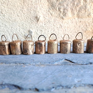 May include: A row of eight small, antique, gold-coloured bells with a loop at the top. The bells are arranged in a line on a grey surface.