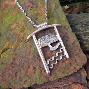 May include: A silver and copper necklace with a pendant featuring a textured, abstract design. The pendant is shaped like a rectangle with a curved top and a wavy bottom. The design includes a crescent moon shape with two smaller circles on top, and a wavy line at the bottom.