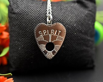 Könnte beinhalten: Eine silberne Halskette mit einem herzförmigen Anhänger, auf dem "SPIRIT" steht. Der Anhänger ist ein Ouija-Brett-Design mit einem Loch in der Mitte. Die Halskette liegt auf einem schwarzen Samtkissen.