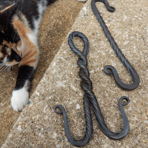 May include: Three black metal hooks with a twisted rope design. The hooks are hanging on a concrete surface.