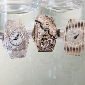 Puede incluir: Tres relojes de pulsera vintage con correas y carátulas de metal plateado. Los relojes están dispuestos en fila sobre una superficie reflectante. El reloj de la izquierda tiene una carátula blanca con números y manecillas negros, y el texto "Bulova" impreso en la carátula. El reloj del medio está abierto, revelando el mecanismo interno del reloj. El reloj de la derecha tiene una carátula blanca con números y manecillas negros, y el texto "Waltham" impreso en la carátula.