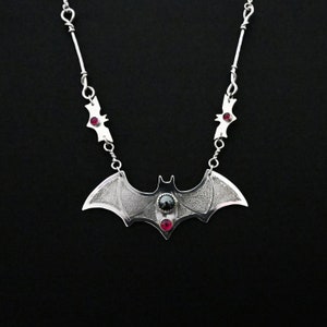 May include: A silver bat necklace with three red gemstones and a black gemstone in the center. The bat is detailed with a textured surface.
