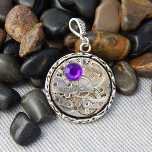 May include: A silver pendant necklace with a round, open-faced watch movement. The watch movement has a purple gemstone in the center and is surrounded by silver gears. The pendant is on a silver chain.