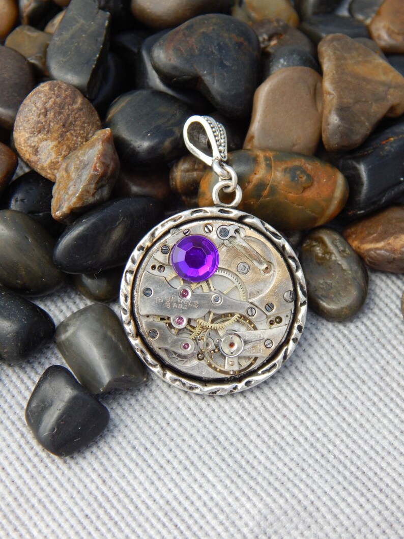 May include: A silver pendant necklace with a round, open-faced watch movement. The watch movement has a purple gemstone in the center and is surrounded by silver gears. The pendant is on a silver chain.
