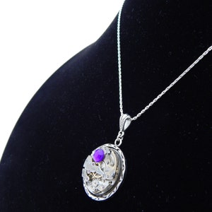 May include: A silver necklace with a pendant featuring a circular clockwork design with a purple gemstone in the center.