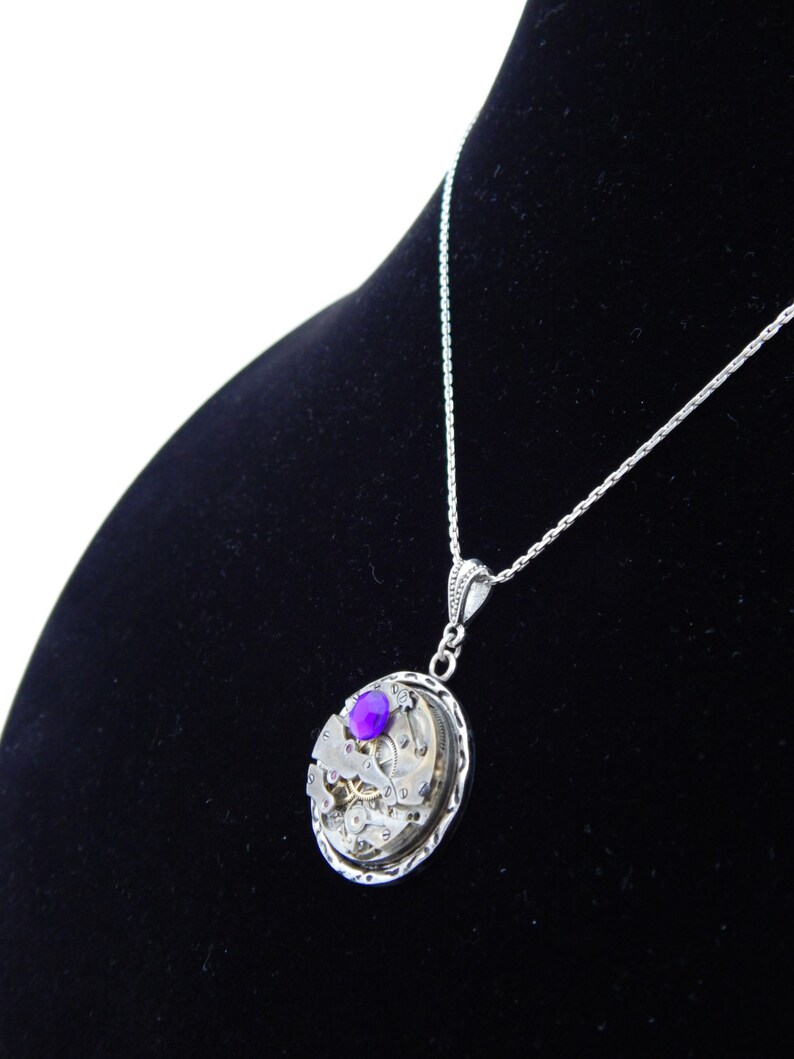 May include: A silver necklace with a pendant featuring a circular clockwork design with a purple gemstone in the center.