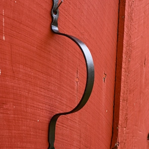 May include: A black metal hook attached to a red wooden door. The hook is shaped like a curved S and has a pointed top.