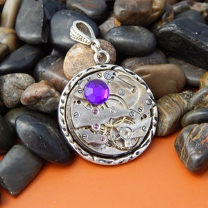 May include: A silver pendant with a purple gemstone set in a circular watch face design. The pendant is surrounded by small silver gears and is on a bed of smooth gray stones.