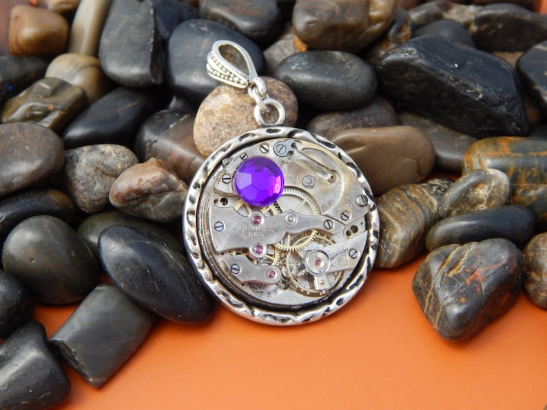 May include: A silver pendant with a purple gemstone set in a circular watch face design. The pendant is surrounded by small silver gears and is on a bed of smooth gray stones.