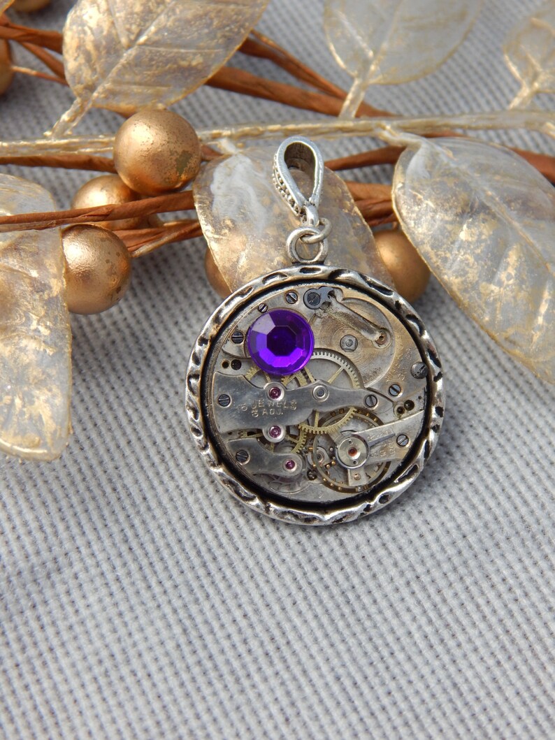 May include: A silver pendant with a purple gemstone set in a circular watch face with visible gears and the text "17 Jewels Swiss Made".