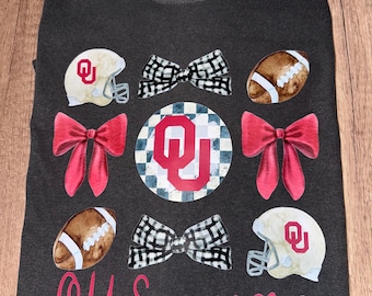May include: A dark gray t-shirt with a red and white OU logo, two footballs, and four bows. The text "OU Sooners" is written in pink.