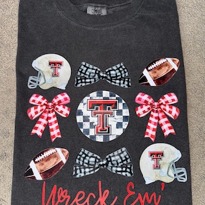May include: A dark gray t-shirt with a red, white, and black design featuring a football helmet, football, and bows. The text "Wreck Em'" is printed in red on the bottom of the shirt.